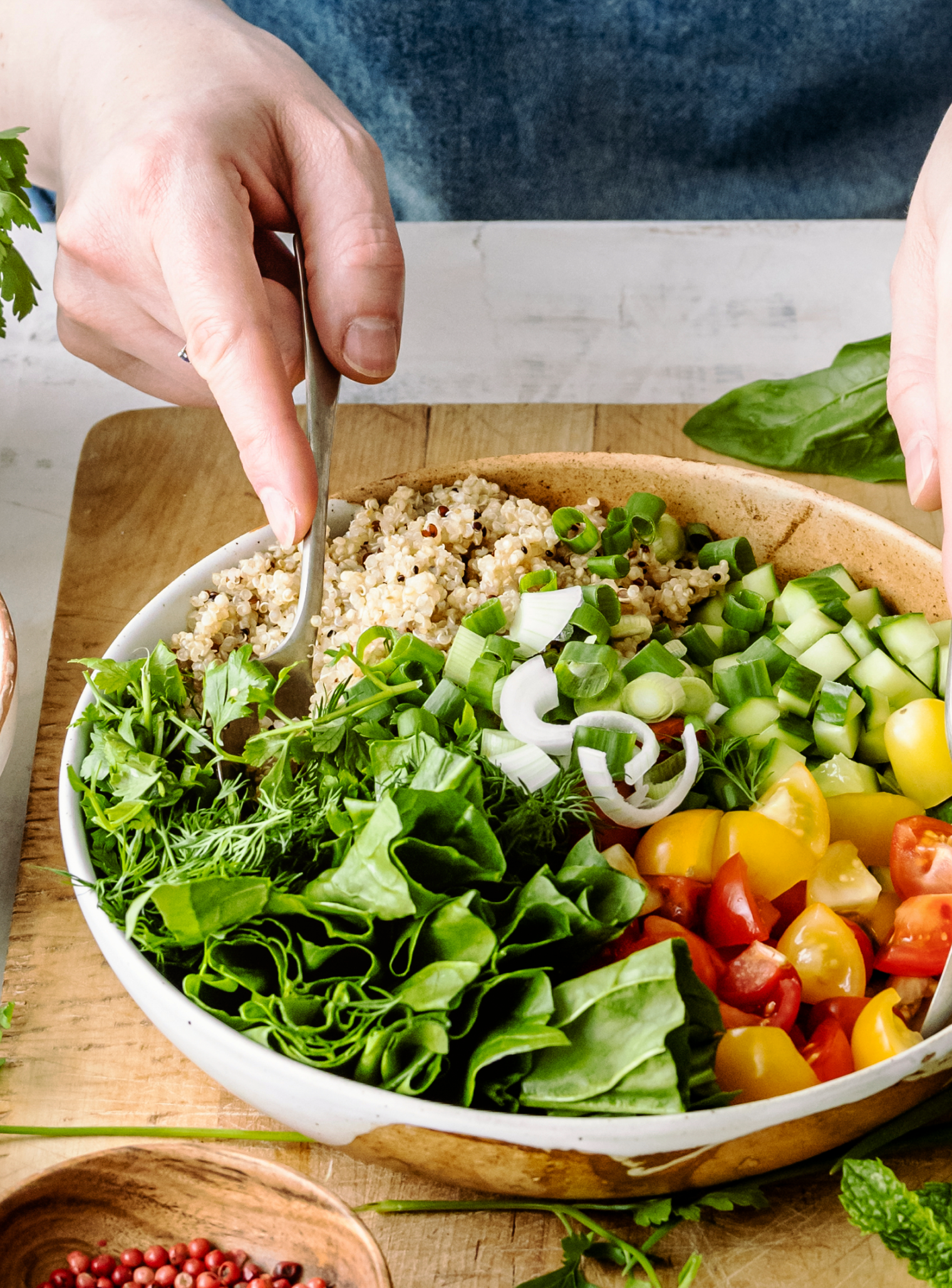 Hand preparing a colorful healthy quinoa salad bowl with fresh vegetables including cucumbers, cherry tomatoes, spinach, parsley, dill, and green onions on a wooden board — holistic nutrition, clean eating, functional wellness lifestyle