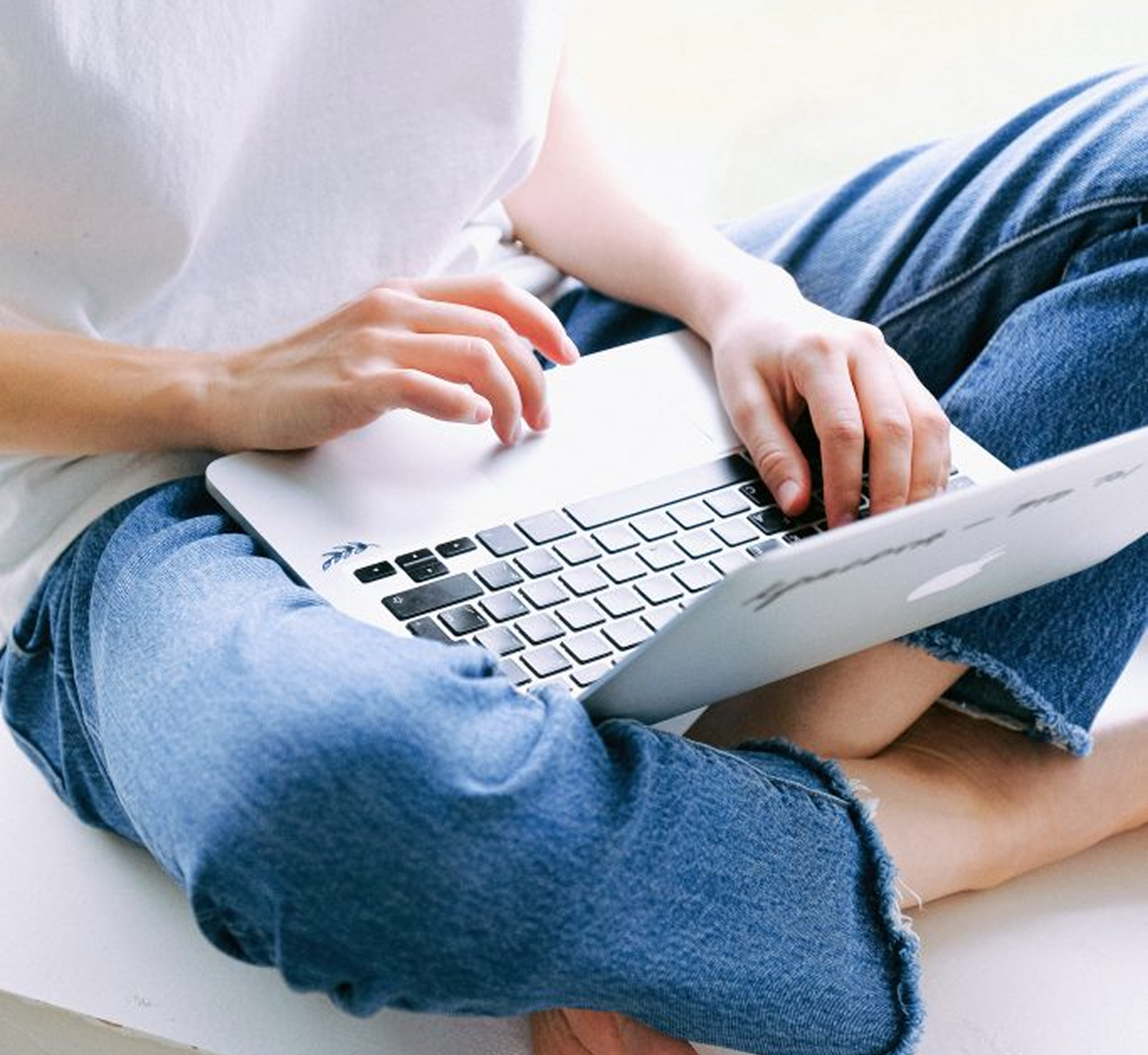 Woman working on a laptop in casual jeans and white t-shirt while sitting cross-legged, representing remote wellness coaching or online nutrition consultation from home