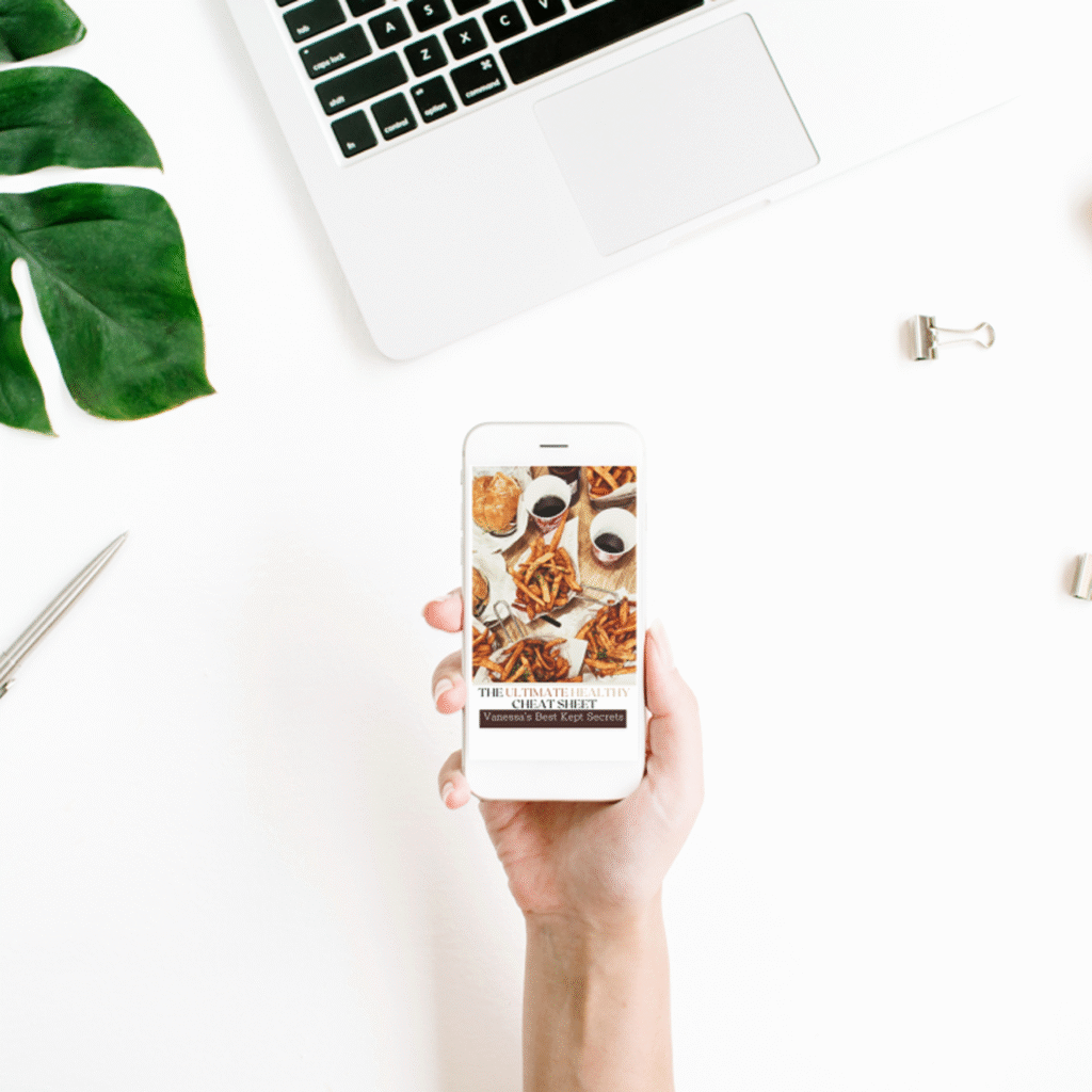 Hand holding phone displaying "The Ultimate Healthy Cheat Sheet" guide, next to laptop and tropical leaf on white desk | digital nutrition ebook | wellness resources | healthy eating tips | dietitian-approved download | Pinnacle Nutrition Group
